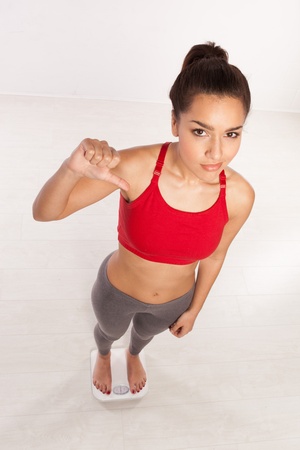 High angle view of an athletic young woman in sportswear standing on a scale giving a thumbs down gesture of disappointment and disapprovalの写真素材