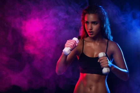 Determined fit sexy woman working out with dumbells on blue and violet smoky wave backgroundの写真素材