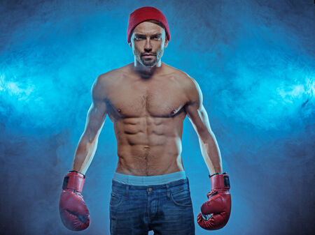 Young athletic boxer exhibiting his six-pack abdominals wearing gloves, blue jeans and a beanie hat...