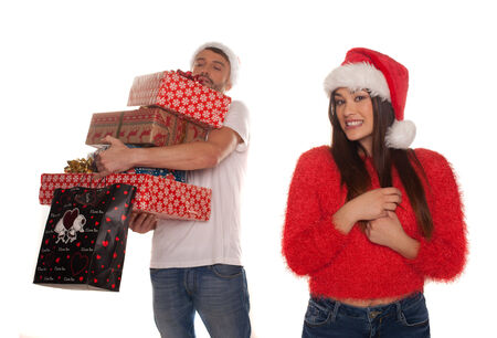 Young couple laden with Christmas presents with the woman standing smiling in the foreground in a festive red Santa Hat while the laughing man holds all the gifts behind her isolated on whiteの写真素材