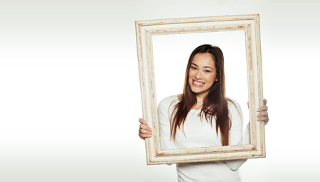 Smiling beautiful young woman holding an old white painted wooden picture frame so that it frames...