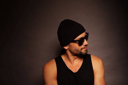 Trendy young man portrait looking away in a dark background wearing a beanie hat, tank top and sunglassesの写真素材