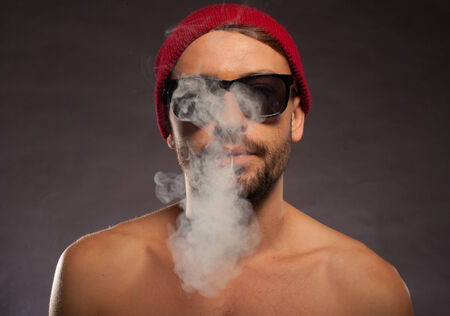 Portrait of a shirtless young man blowing cigarette smoke in a dark backgroundの写真素材