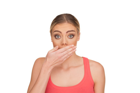 Portrait of a surprised blond woman covering the mouth with the hand in disbelief and eyes wide open wearing a red tank top on a white backgroundの写真素材