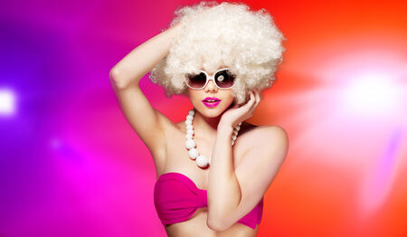 Glamorous woman with a blond afro hairstyle and trendy sunglasses and necklace posing against a colourful background with light flaresの写真素材