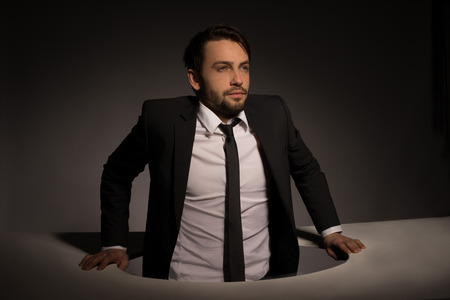 Handsome young bearded businessman climbing out of a circular hole in a white wall in a conceptual imageの写真素材