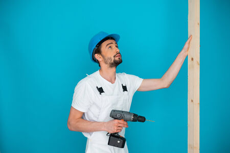 Workman in a hardhat holding a battery-operated hand drill and a plank of wood as he does carpentry on a construction site or during home renovations, on a blue wallの写真素材