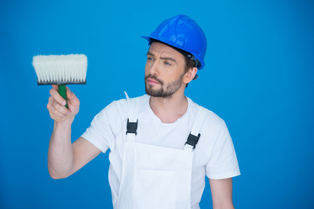 Attractive bearded young painter in dungarees standing looking at the camera holding up a white paint brush on a bluish backgroundの写真素材