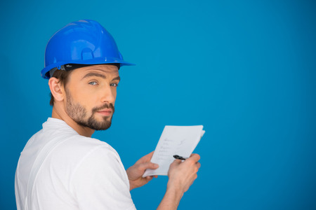 Artisan or workman taking notes standing in front of a blue wall with a pen and paper in his hands turning to look at the camera with a serious thoughtful expressionの写真素材