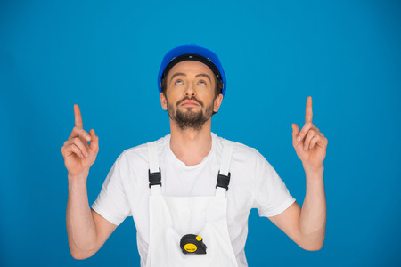 Handsome young male workman in a hardhat pointing above his head with both hands as he looks up with his head tilted back, blue backgroundの写真素材