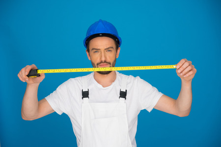 Young builder or workman in a hardhat and dungarees holding up a measuring tape extended in front of...