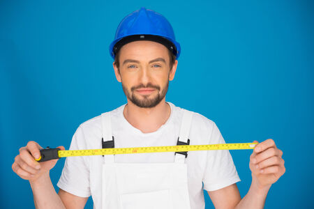 Smiling young workman wearing a hardhat and dungarees standing holding a tape measure extended in his hands against a blue backgroundの写真素材