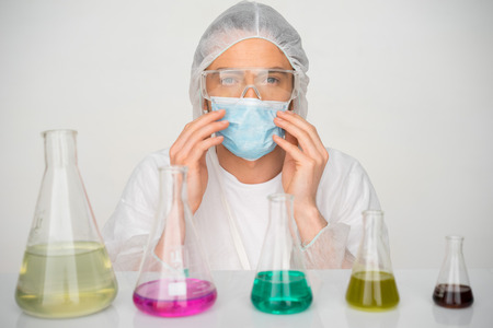Male technician wearing a mask and cap working in a chemistry laboratory sitting behind a row of...