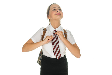 Proud young schoolgirl in school uniform standing with her backpack over her shoulder straightening her tie with a smile, on whiteの写真素材