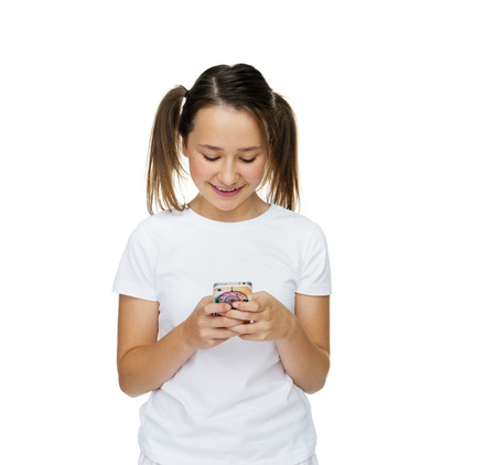 Young girl with her hair in pigtails standing reading a text message on her mobile phone smiling with pleasure at the news, on whiteの写真素材