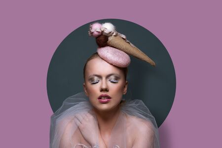 Fine art portrait of a gorgeous woman in fascinator hat in the shape of an ice cream cone with creative makeup and filmy gauze around her shoulders over a purple backgroundの写真素材