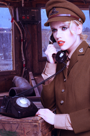 Pretty blond woman in a brown army uniform standing talking on a retro dialup telephone on a wooden crate in a barracksの写真素材