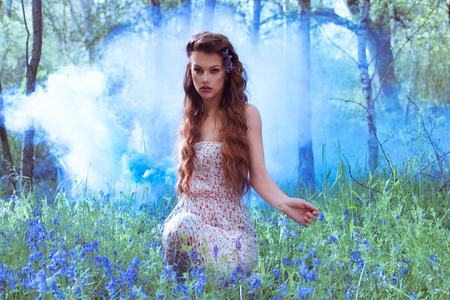 Artistic portrait of a pretty young redhead girl in a bluebell forest kneeling amongst the flowers with an applied blue filter for ambiance and atmosphereの写真素材