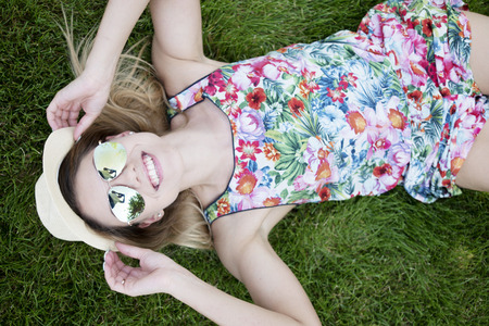 Pretty Young Woman in a Trendy Outfit Smiling at the Camera From High Angle View While Lying on the Green Grasses and Holding her Sunglasses.の写真素材