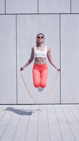 Energetic Young Woman in Fitness Outfit, Exercising Using Jumping Rope Against White Wall Background...
