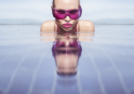 Face closeup of sexy young woman wearing purple sunglasses looking at camera in infinity rooftop swimming pool on a sunny day over blue sky and green trees landscapeの写真素材