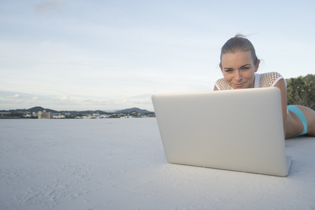 Outdoors portrait of pretty blonde woman in bikini with laptop computer lying on a rooftop over city landscape and sky background. Freelance and technologyの写真素材