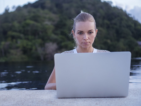 Outdoors portrait of pretty blonde woman in white top with laptop computer standing in a rooftop swimming pool over blurred green mountain and sky background. Freelance and technologyの写真素材