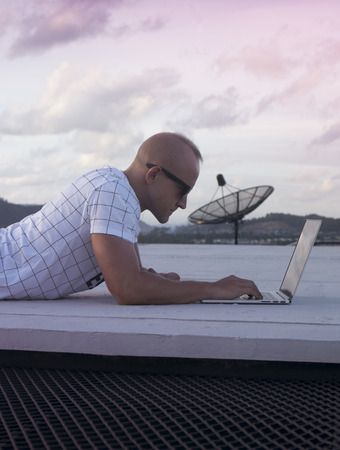 Outdoors portrait of handsome young man in sunglasses with laptop computer lying on a rooftop over sunset sky background. Freelance and technologyの写真素材