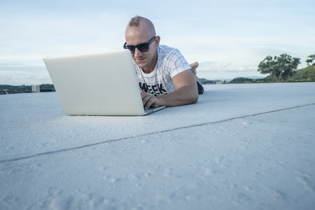 Outdoors portrait of handsome young man in sunglasses with laptop computer lying on a rooftop over sky background. Freelance and technologyの写真素材