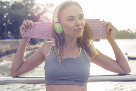 Young pretty blonde fitness female wearing bright green headphones listening music and holding pink skateboard while standing on the pier near the sea, enjoying early morning with beautiful sunriseの写真素材