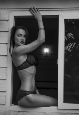 Black and white photo of young beautiful sexy woman in lingerie sitting and looking out of window and enjoying morning sunlight. Seductive look of girl admiring shining sunの写真素材