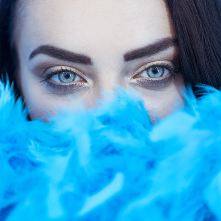 Closeup portrait of beautiful seductive brunette woman smiling and looking into the camera while playing with blue feather boa over blue wall backgroundの写真素材