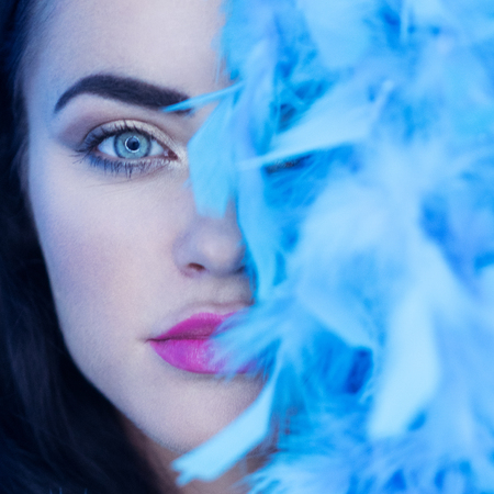 Closeup portrait of beautiful seductive brunette woman smiling and looking into the camera while playing with blue feather boa over blue wall backgroundの写真素材