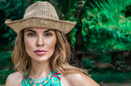 Portrait of beautiful young woman with straw hat on a sunny day looking into the camera over palm trees background. Beautiful eyes closeup of pretty girl wearing colorful bikini top and straw hat over greenの写真素材