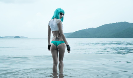 Sexy beautiful woman in modern futuristic style posing in the sea. Creative look of woman wearing bikini, blue wig, black leather fingerless gloves in the waterの写真素材