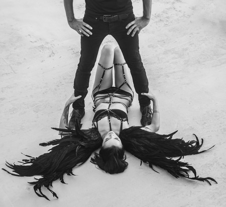 Black and white photo of romantic scene with man's silhouette standing above sensual angel woman wearing lingerie, leather belts and high heels lying on the floorの写真素材