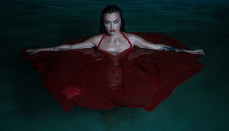 Beautiful mysterious woman looking like a vampire wearing red dress looking into the camera ehile standing in the pool at nightの写真素材