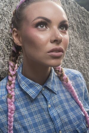 Portrait of young pretty girl with pink plaits wearing blue checked shirt sitting on the rock during summer dayの写真素材