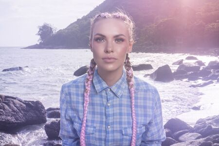 Portrait of young pretty girl with pink plaits wearing blue checked shirt looking into the camera while sitting on the rock during summer day over sea and sky backgroundの写真素材