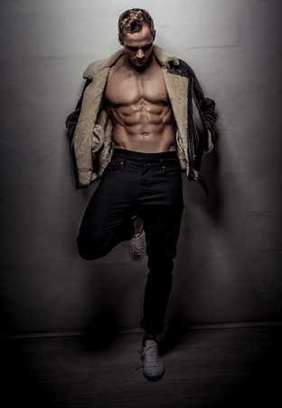 Attractive muscular athlete young man wearing pants and leather jacket looking down while leaning on wall isolated on light gray backgroundの写真素材