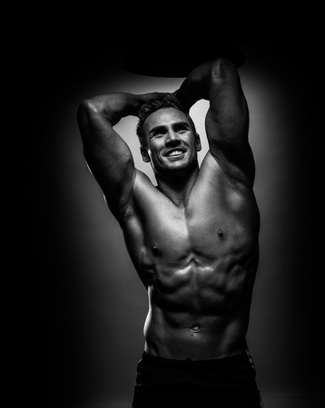Attractive shirtless muscular athlete young man smiling while standing with hands on head in black and white studio, vertical photoの写真素材