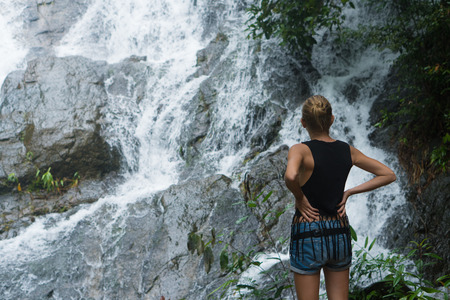 Back view of blonde woman standing on a rock and looking up near waterfall. Female tourist enjoying by a water fall in forestの写真素材