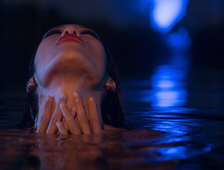 Closeup face of sexy Asian woman with wet hair standing in the swimming pool with head lying down on the water and hands on her neck during summer evening over blue lights backgroundの写真素材