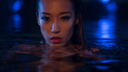 Closeup face of sexy Asian woman with wet hair standing in the swimming pool and looking into the camera during summer evening over blue lights backgroundの写真素材