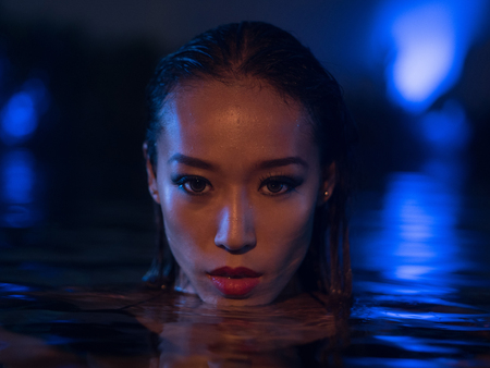 Closeup face of sexy Asian woman with wet hair standing in the swimming pool and looking into the camera during summer evening over blue lights backgroundの写真素材