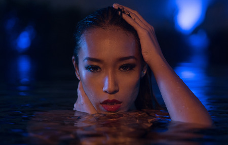 Closeup face of sexy Asian woman with wet hair standing in the swimming pool and looking into the camera during summer evening over blue lights backgroundの写真素材