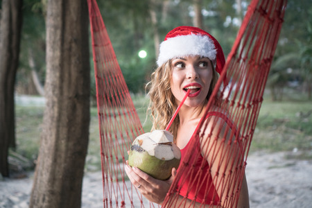 Young happy woman in santa hat and red swimsuit sitting on the hammock and drinking from coconut on the sandy beach. Christmas beach vacation travel woman enjoying christmas on tropical beachの写真素材