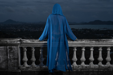 Back view of mysterious woman in black bikini and blue hoody cover up sitting on old stone ornate carved balustrade over cloudy sky and cityscapeの写真素材