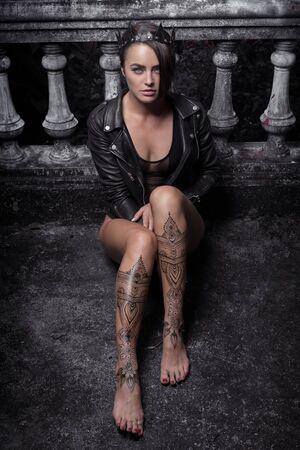 Mysterious beautiful woman in black bodysuit, leather jacket and crown with henna tattoo on her legs sitting leaning against old stone steps with ornate carved balustrade while looking at the cameraの写真素材