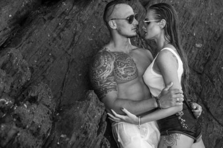 Sensual young couple in wet clothes and sunglasses looking at each others while leaning against the rock by the sea - black and white photoの写真素材